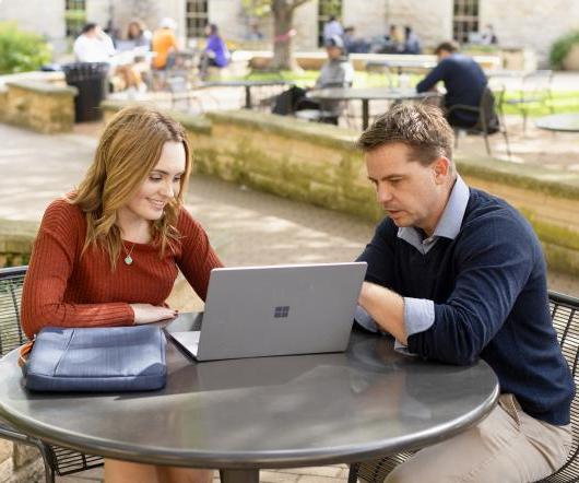 A professor and a student sit at a patio table and look at a laptop.